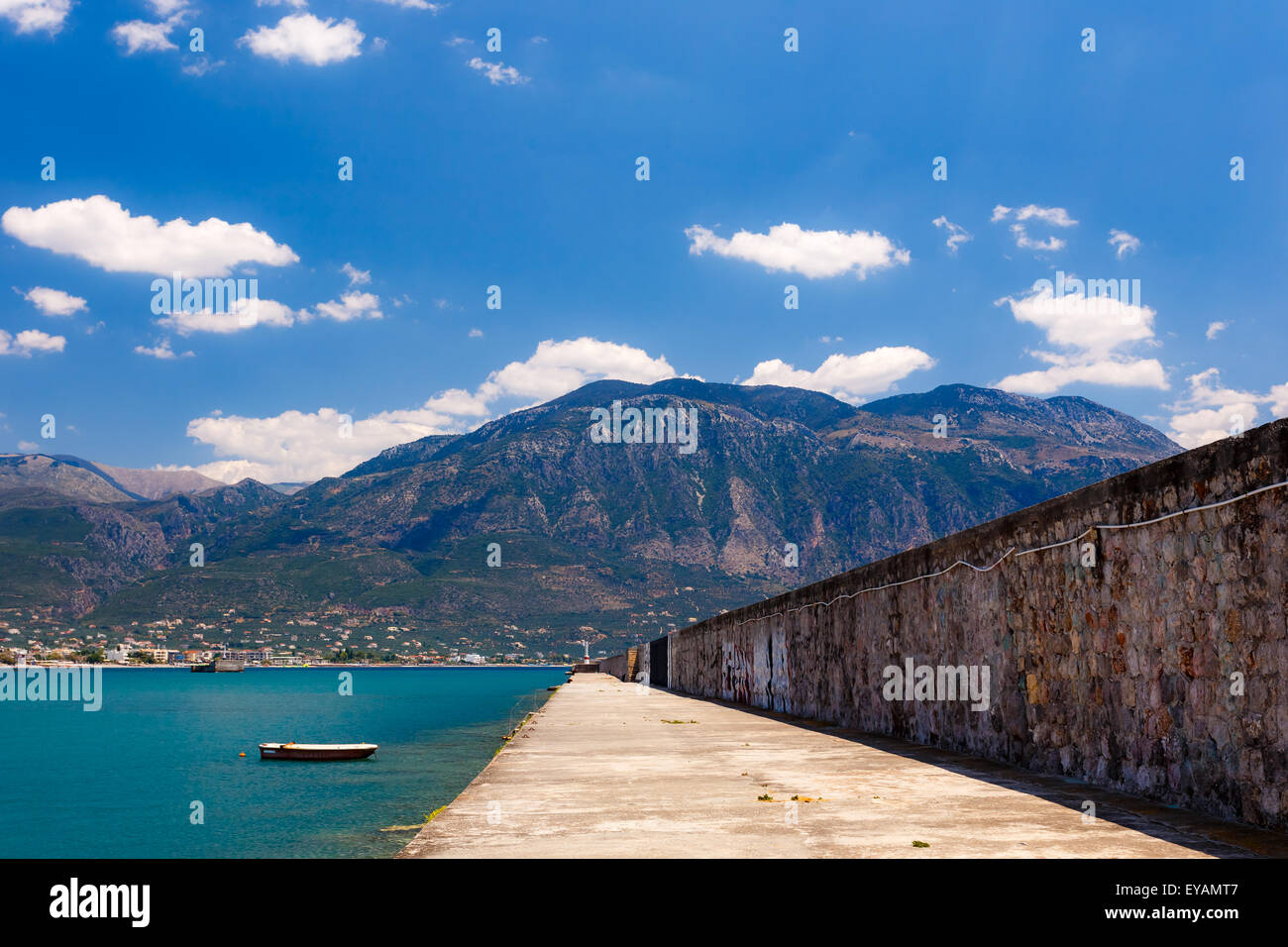 I pesci di piccola barca ancorata in porto con piccole città sullo sfondo e la lunga strada laterale contro un cielo blu e nuvole in Grecia Foto Stock