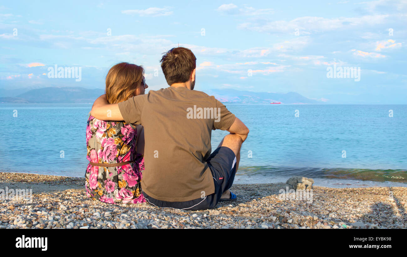 Giovani amare giovane seduti insieme su una spiaggia rocciosa e guardando il mare Foto Stock