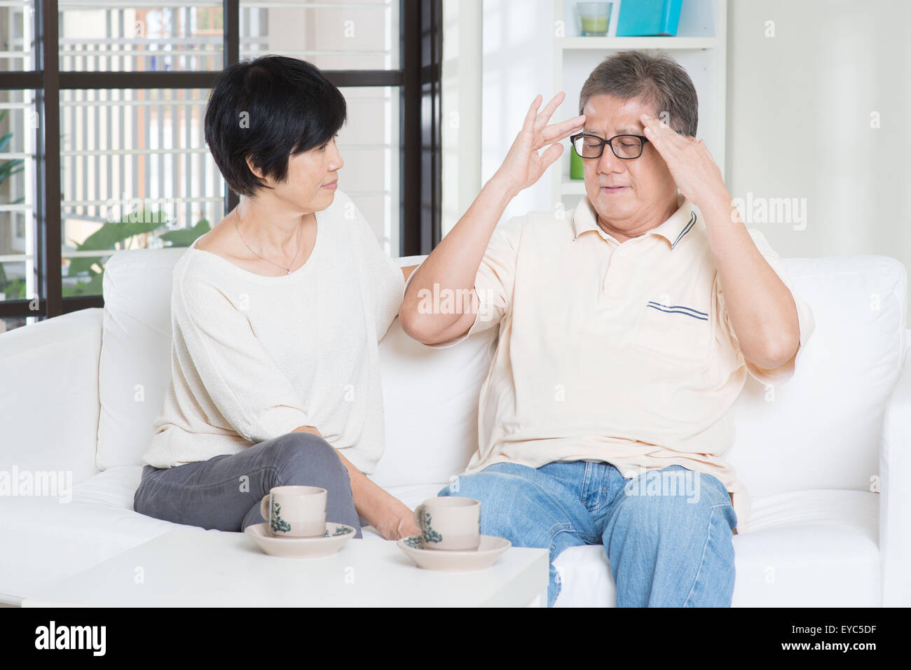 Asian vecchio uomo mal di testa, seduto sul divano con mia moglie a casa. Famiglia cinese, senior pensionato in interni vivere lo stile di vita. Foto Stock