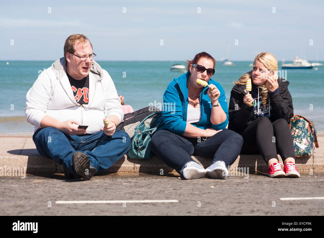 Tre persone sedute su un marciapiede mangiare ghiaccioli a Swanage in luglio Foto Stock