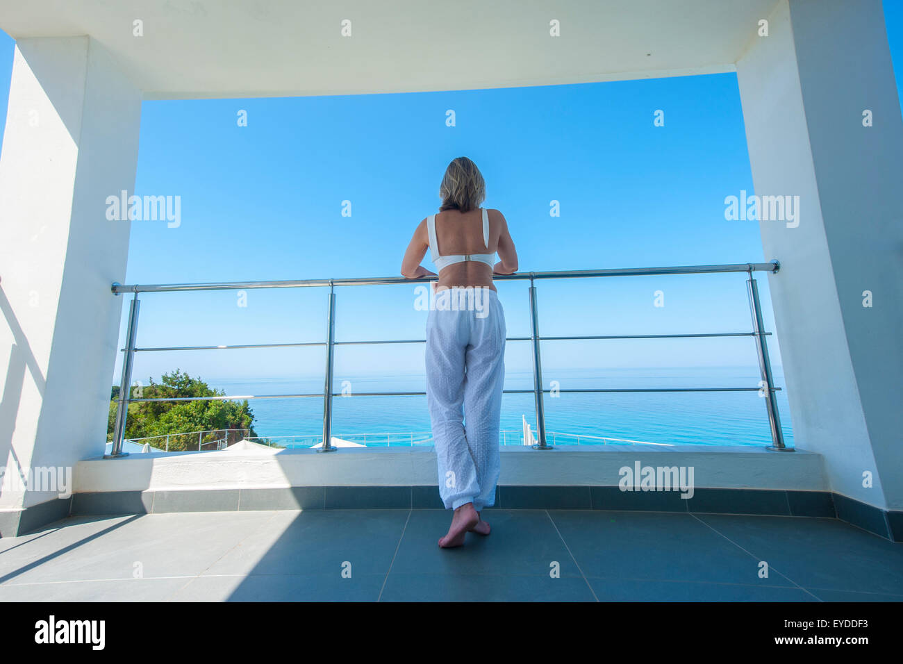 Una dama elegante sorge sul balcone di una villa di lusso in la mattina presto che si affaccia su un mare azzurro Foto Stock