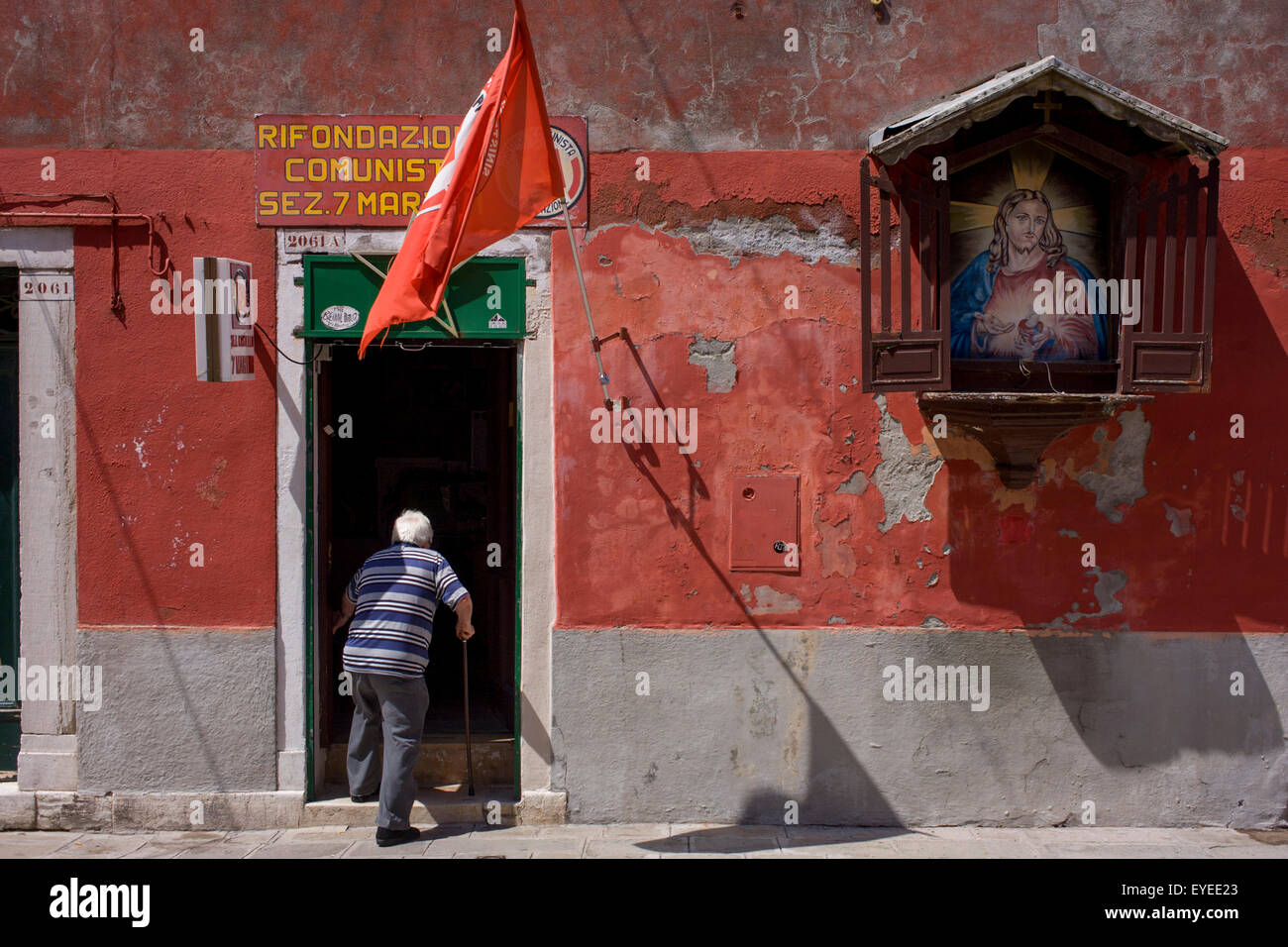 Un uomo anziano con un bastone da passeggio entra le ombre del partito comunista italiano in ufficio con una strada Santuario di Gesù sulla sua parete, Foto Stock