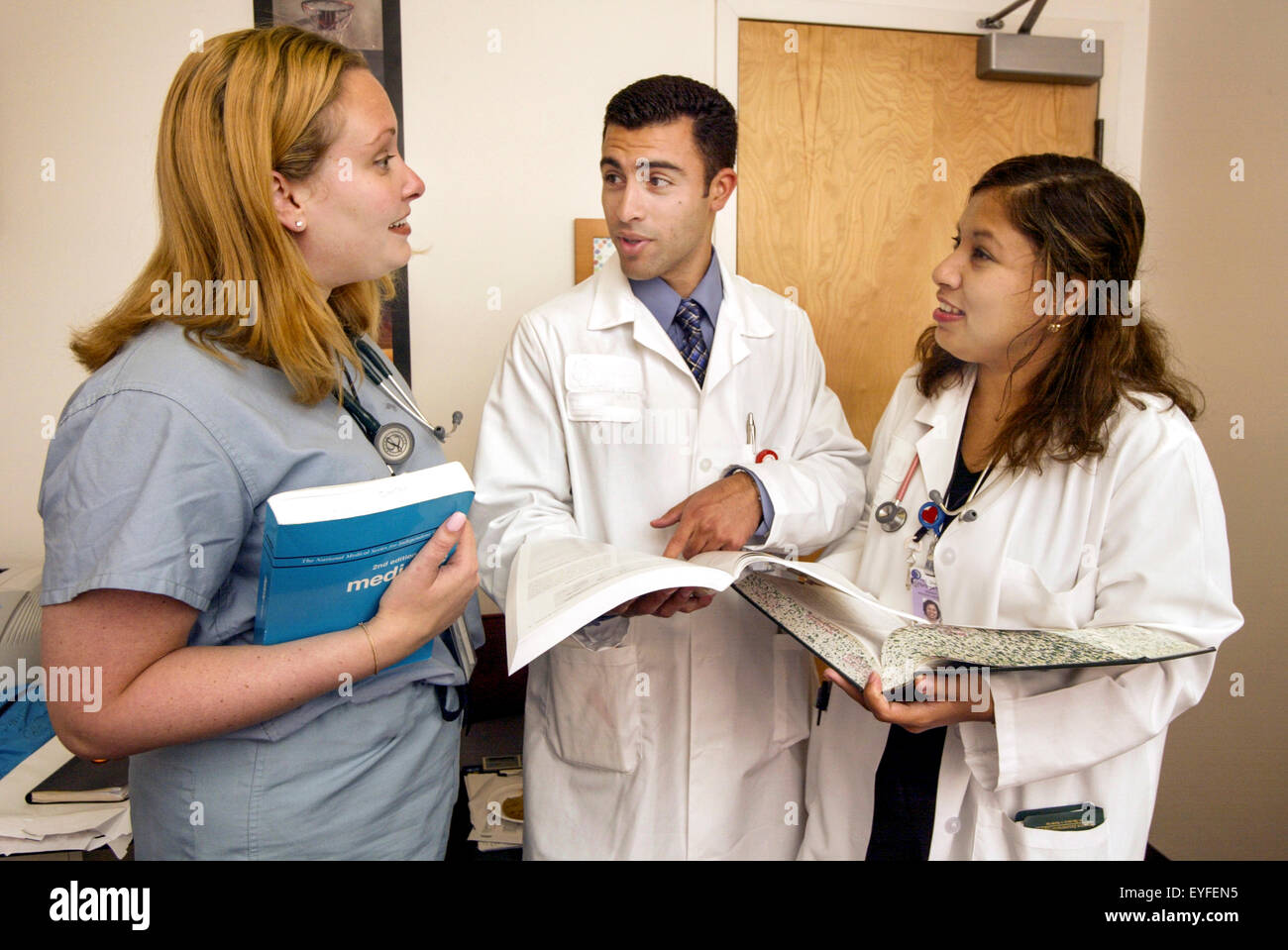 Tre multirazziale medici residenti presso un Orange, CA, ospedale consultare libri di riferimento e discutere le loro scoperte. Nota camici e frega. Foto Stock
