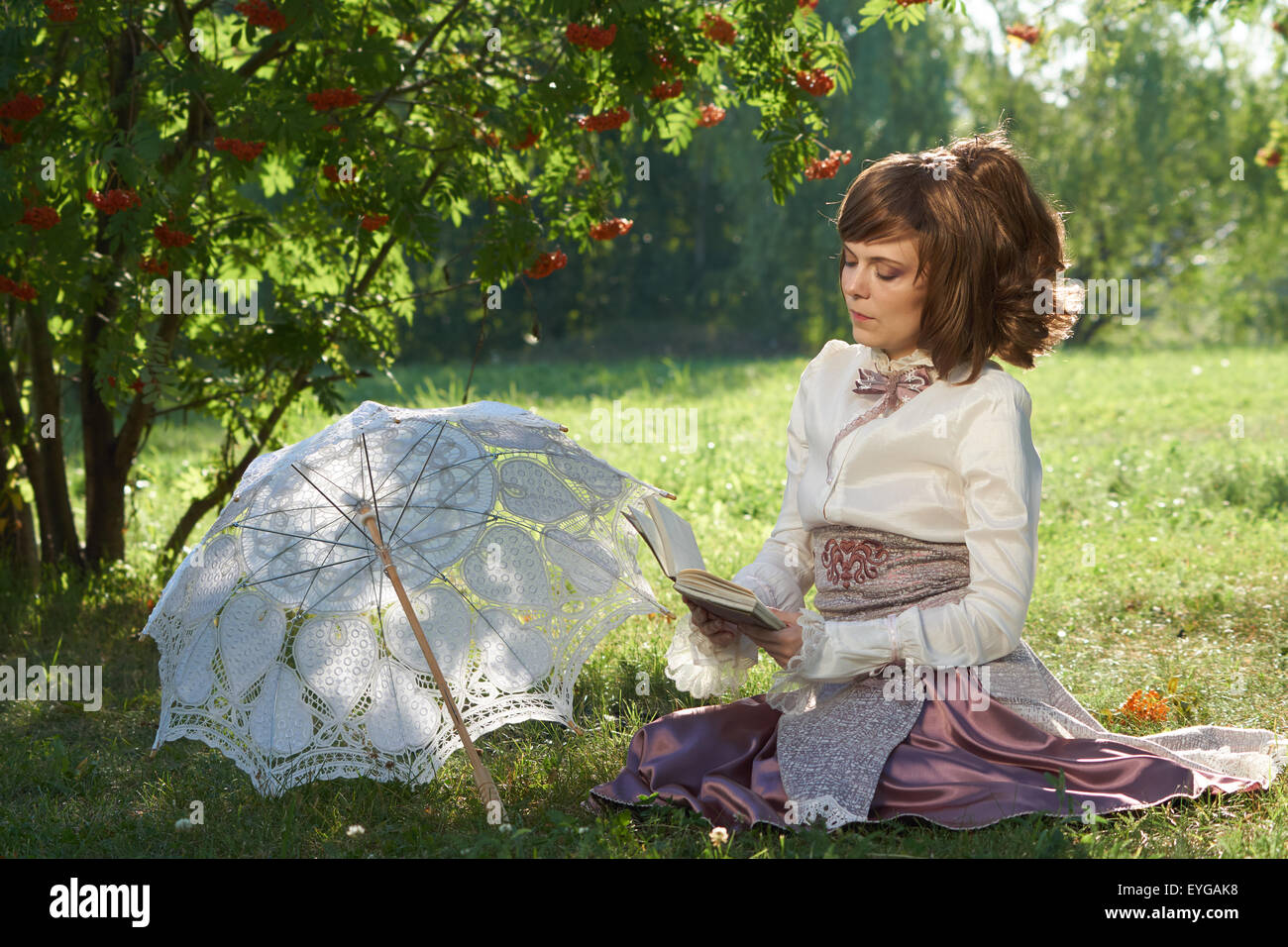 Bellissima ragazza si siede sul prato del parco estivo sotto rowan tree e legge libri interessanti con il bianco ombrellone accanto Foto Stock