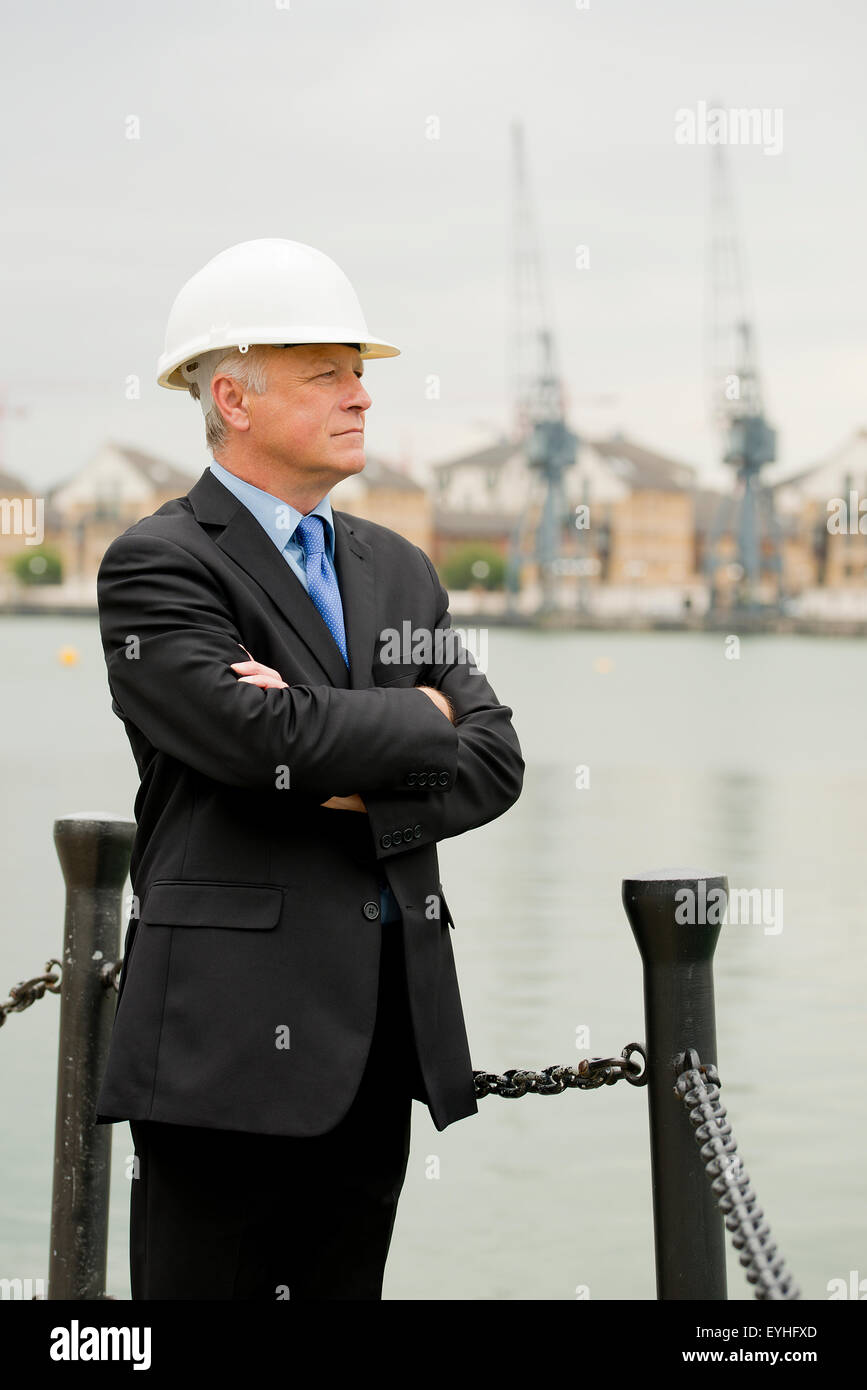 Imprenditore che indossa un abito scuro e bianco casco di sicurezza, sorge con le sue braccia piegate in parte anteriore del dock. Foto Stock