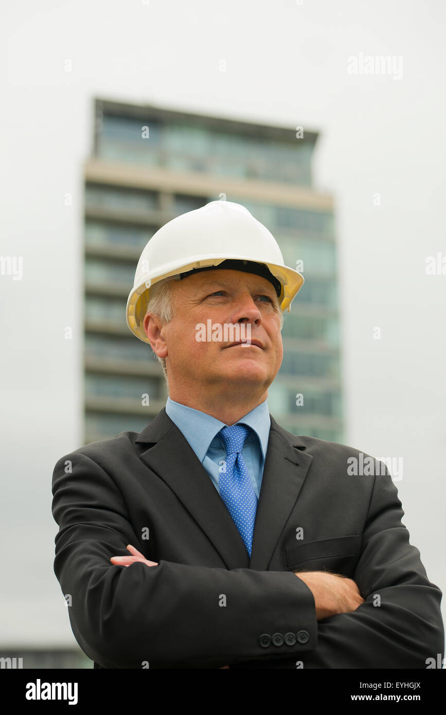 Imprenditore che indossa un abito scuro e bianco casco di sicurezza, sorge con le sue braccia piegate. Foto Stock