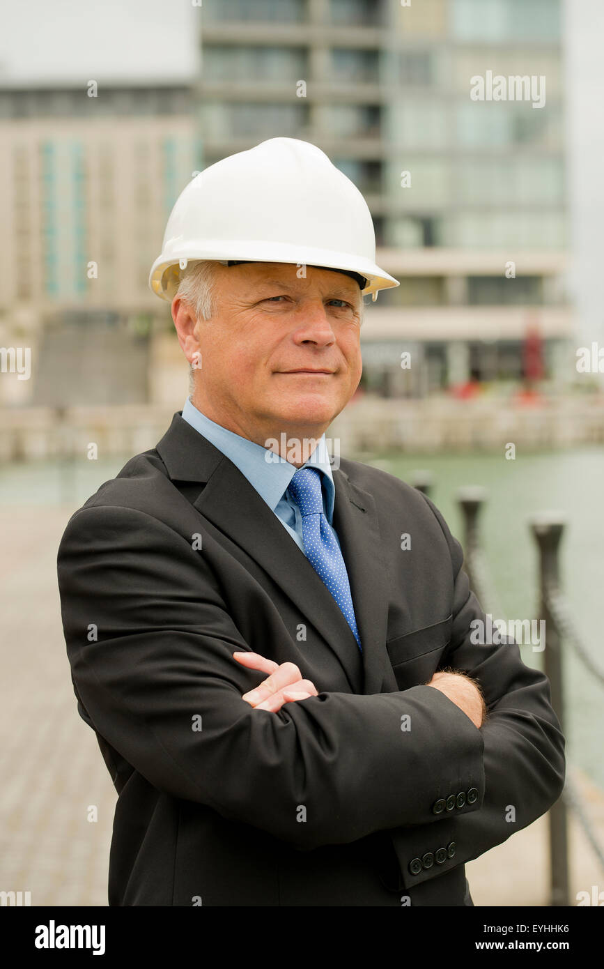 Imprenditore che indossa un abito scuro e bianco casco di sicurezza, sorge con le sue braccia piegate, guardando la fotocamera. Foto Stock