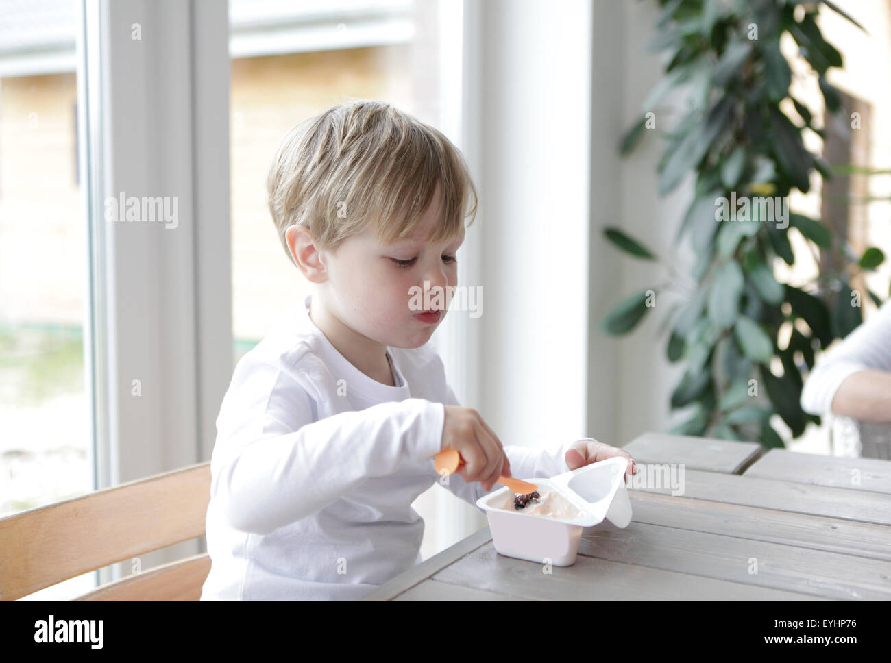 Un piccolo ragazzo seduto al tavolo della cucina, mangiare lo yogurt con un cucchiaio Foto Stock