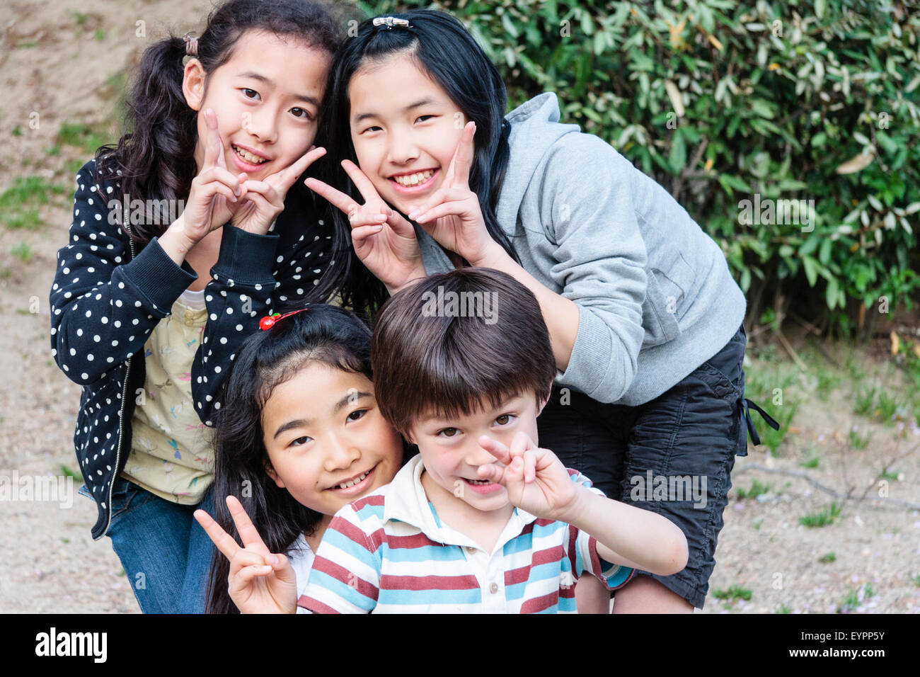 Fila di quattro bambini sorridenti, 7-9 anni, rivolta verso il viewer con contatto visivo. 3 ragazze giapponesi e un ragazzo caucasico tutti di dare pace giapponese gesto. Foto Stock