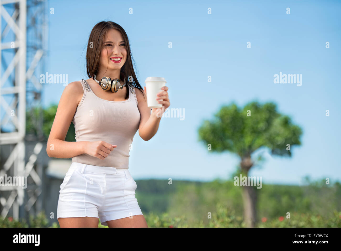Felice giovane donna indossa vintage cuffie musicali intorno al suo collo, azienda di caffè da asporto e in posa sullo sfondo del par Foto Stock