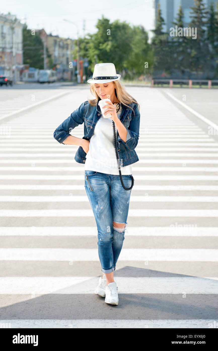 Bella giovane donna in un cappello e una giacca di jeans con un dispositivo  monouso tazza da caffè, in piedi sulla strada con strisce pedonali, bere  Foto stock - Alamy