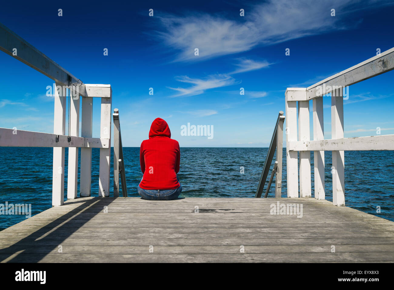 Da solo giovane donna in rosso Maglia con cappuccio seduta sul bordo del molo in legno guardando l'acqua - Disperazione, solitudine, alienazione C Foto Stock