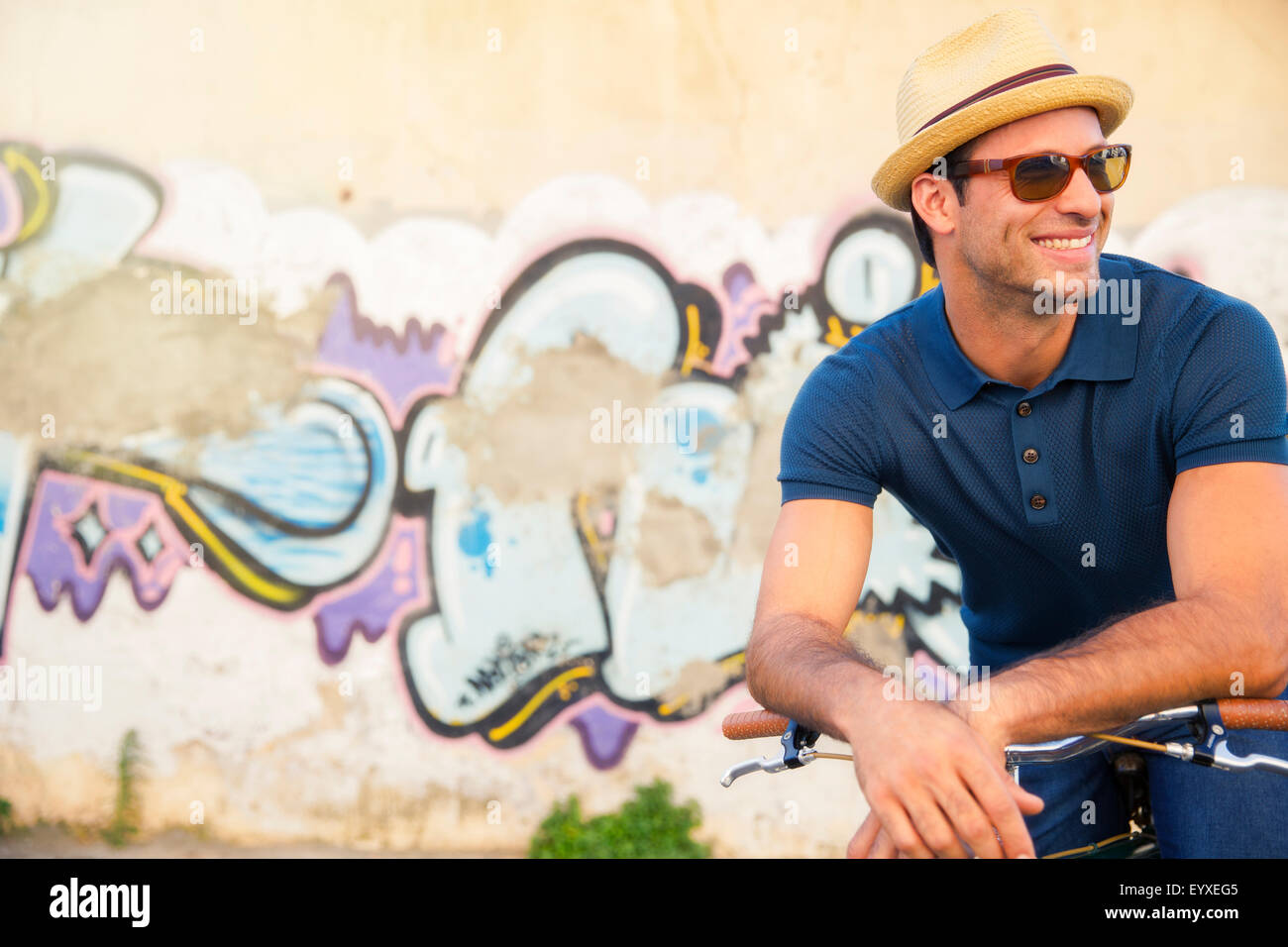 Uomo sorridente in cappello e occhiali da sole appoggiata sulla bicicletta accanto a urban graffiti wall Foto Stock
