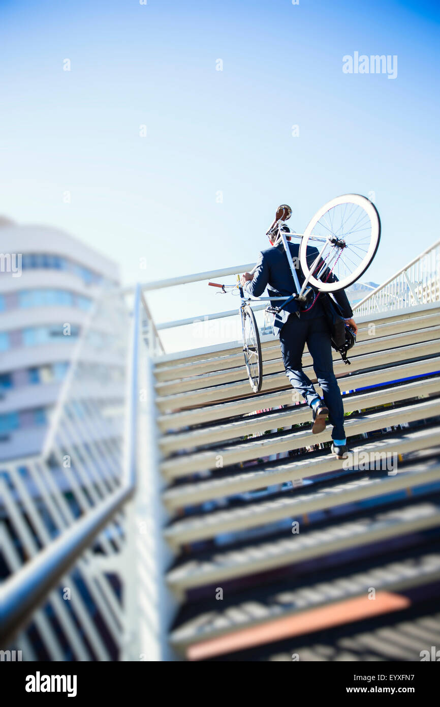 Imprenditore che porta bici fino urban scale sotto sunny blue sky Foto Stock