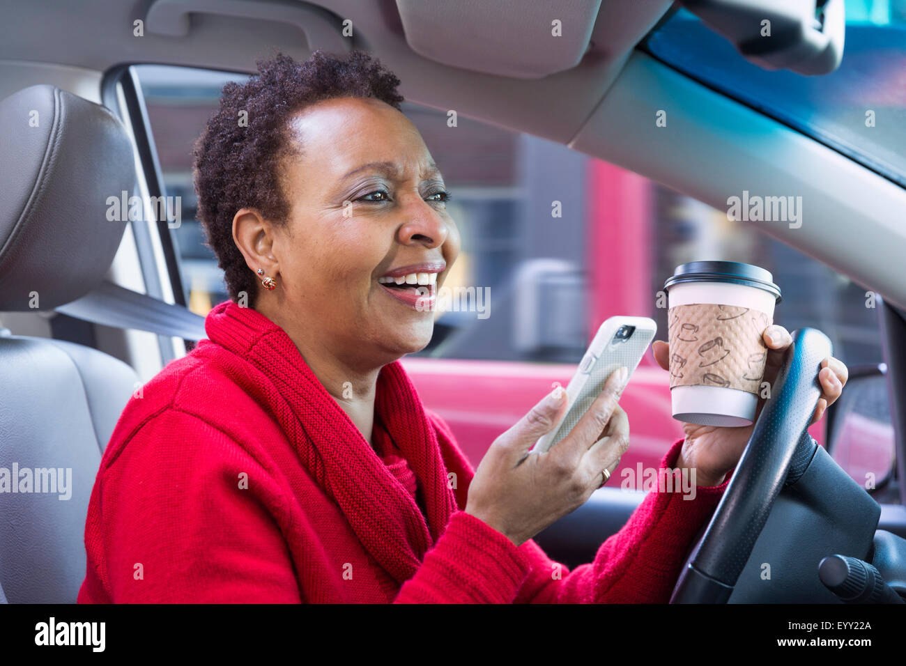Nero donna tramite telefono cellulare e di bere il caffè mentre si guida auto Foto Stock