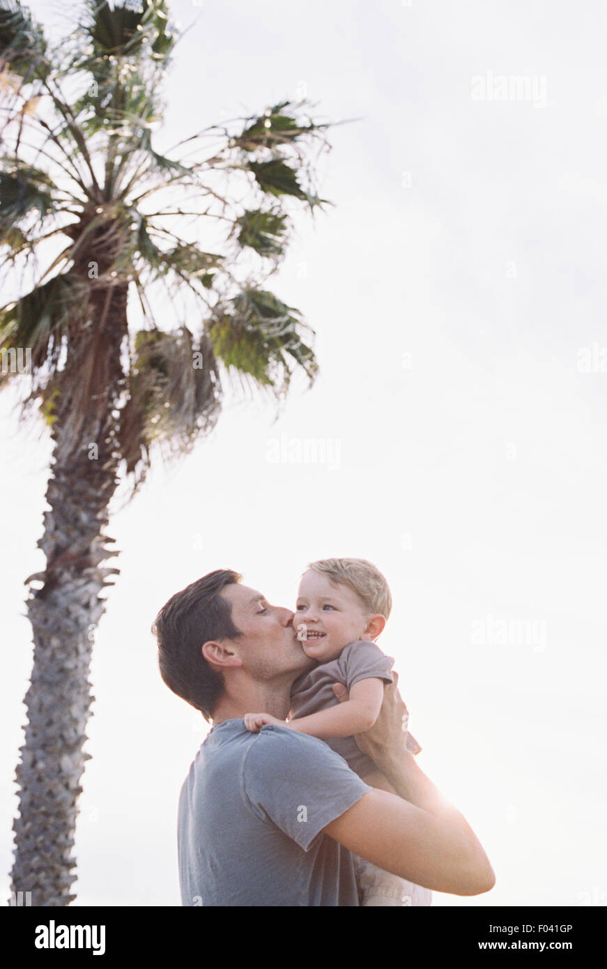 Uomo in piedi da un albero di palma, portando il suo giovane figlio nelle sue braccia, kissing lui sulla guancia. Foto Stock