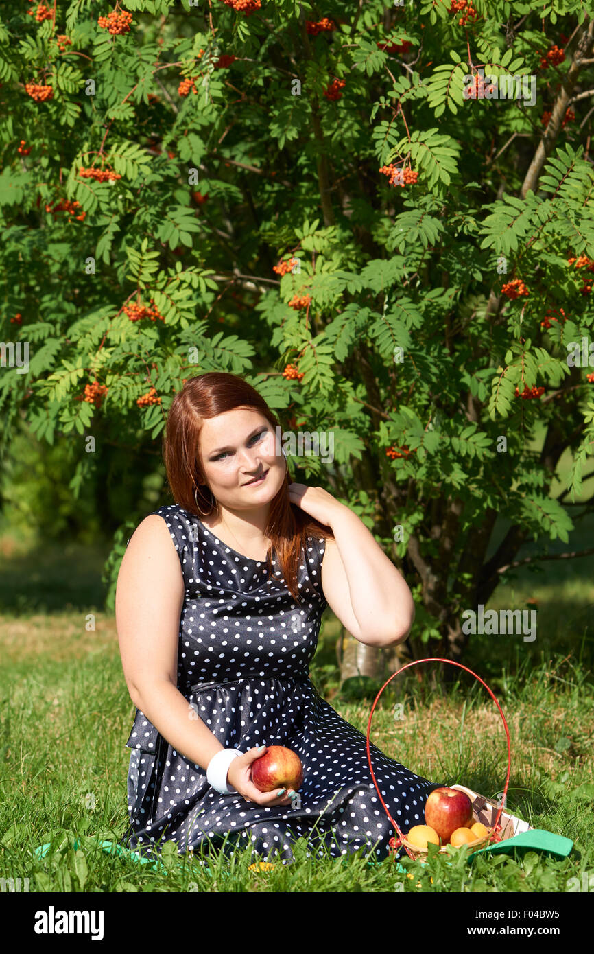 Una ragazza con i capelli rossi e Apple in mano si siede nel prato dell'estate parco e dispone di picnic Foto Stock