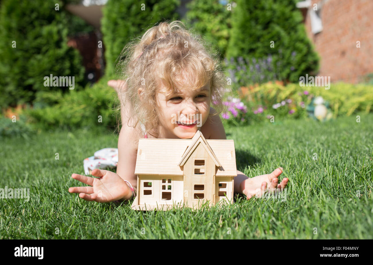Ritratto di bambina giacente su erba con modello in legno di casa in mani, estate all'aperto, un nuovo concetto di home Foto Stock