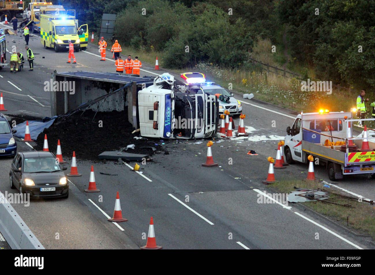 M3, Farnborough, Hampshire, Regno Unito. 10 Agosto, 2015. M3 vicino a Farnborough mostra GV della scena dopo un automezzo entra in collisione con una manutenzione autostrade veicolo parcheggiato sul disco spalla. Il carrello è stato dare protezione ai camion di recupero che stava recuperando un ripartiti automobilista. Il HGV dopo la alta velocità di collisione è venuto a poggiare sul suo lato dopo versando il suo carico su corsie uno e due del carrello principale modo. Due persone sono state portate in ospedale, uno da Air Ambulance la seconda da strada. Credito: Jason Kay/Alamy Live News Foto Stock