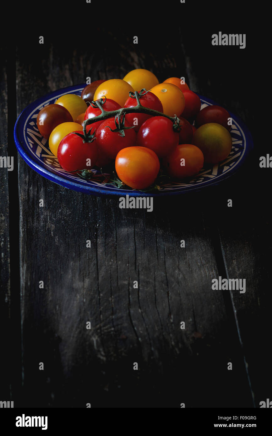 Assortimento di pomodori ciliegia rosso, giallo e arancione nel blu piastra ornamentali su un vecchio tavolo di legno. Scuro in stile rustico. Giorno paglierino Foto Stock
