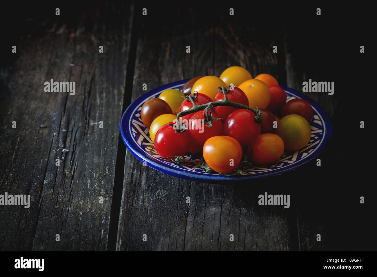 Assortimento di pomodori ciliegia rosso, giallo e arancione nel blu piastra ornamentali su un vecchio tavolo di legno. Scuro in stile rustico. Giorno paglierino Foto Stock