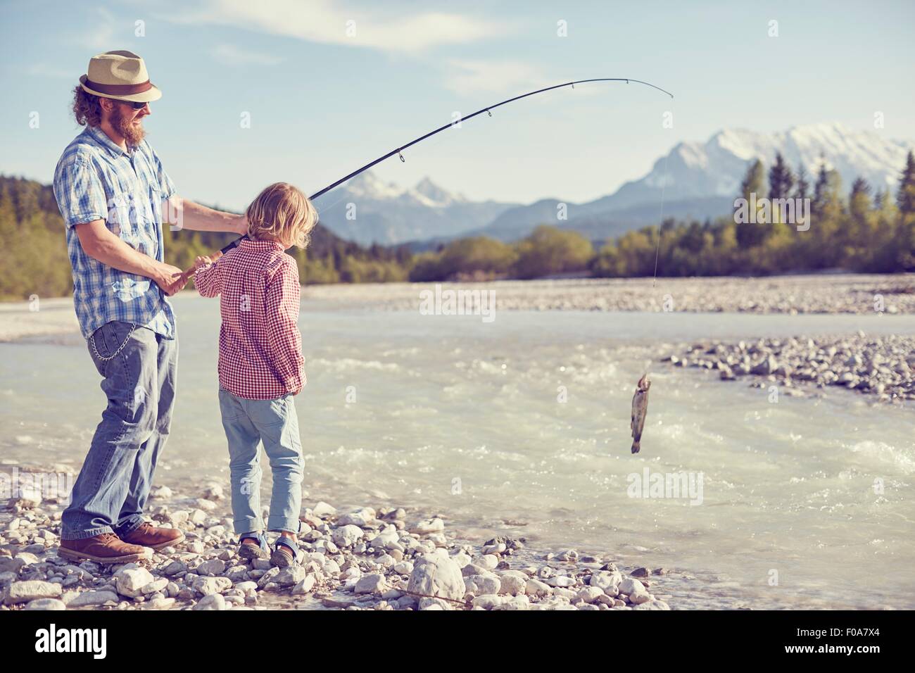 Metà uomo adulto e bambino vicino al fiume tenendo la canna da pesca con pesce attaccato, Wallgau, Baviera, Germania Foto Stock