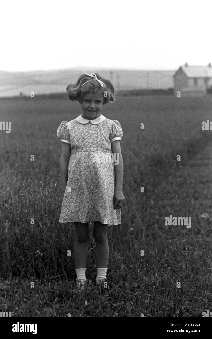 Una bambina indossa un abito, stando in piedi in un campo con la sua famiglia a casa in distanza. Foto Stock