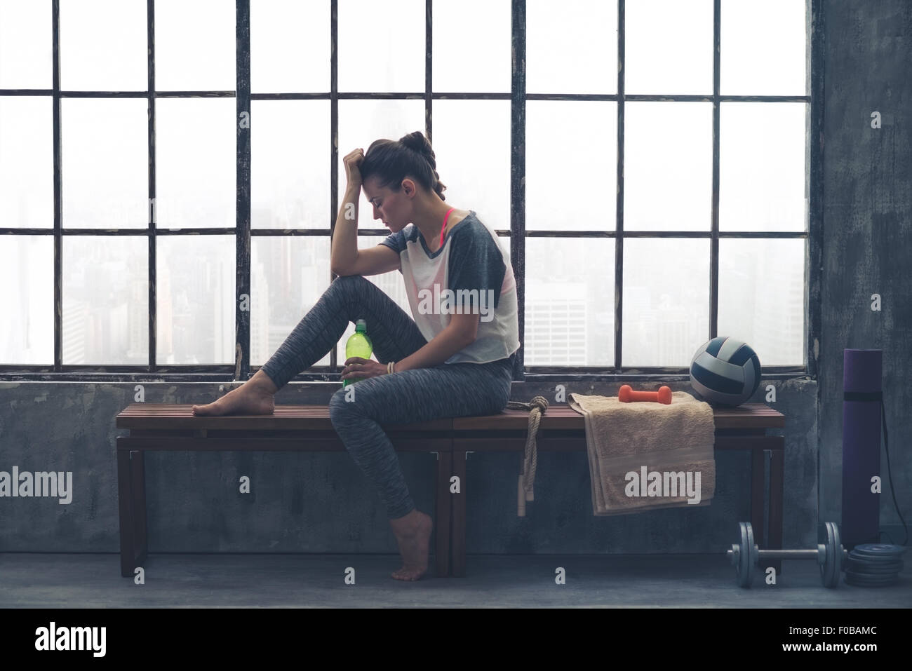 Una donna è seduta su una panca in legno, la gamba in alto, ginocchio piegato ed appoggiando il suo gomito sul suo ginocchio. La sua testa è appoggiata contro la sua mano. Lei è nel profondo del pensiero, stanchi. Nella sua altra, ella mantiene l'acqua. Foto Stock