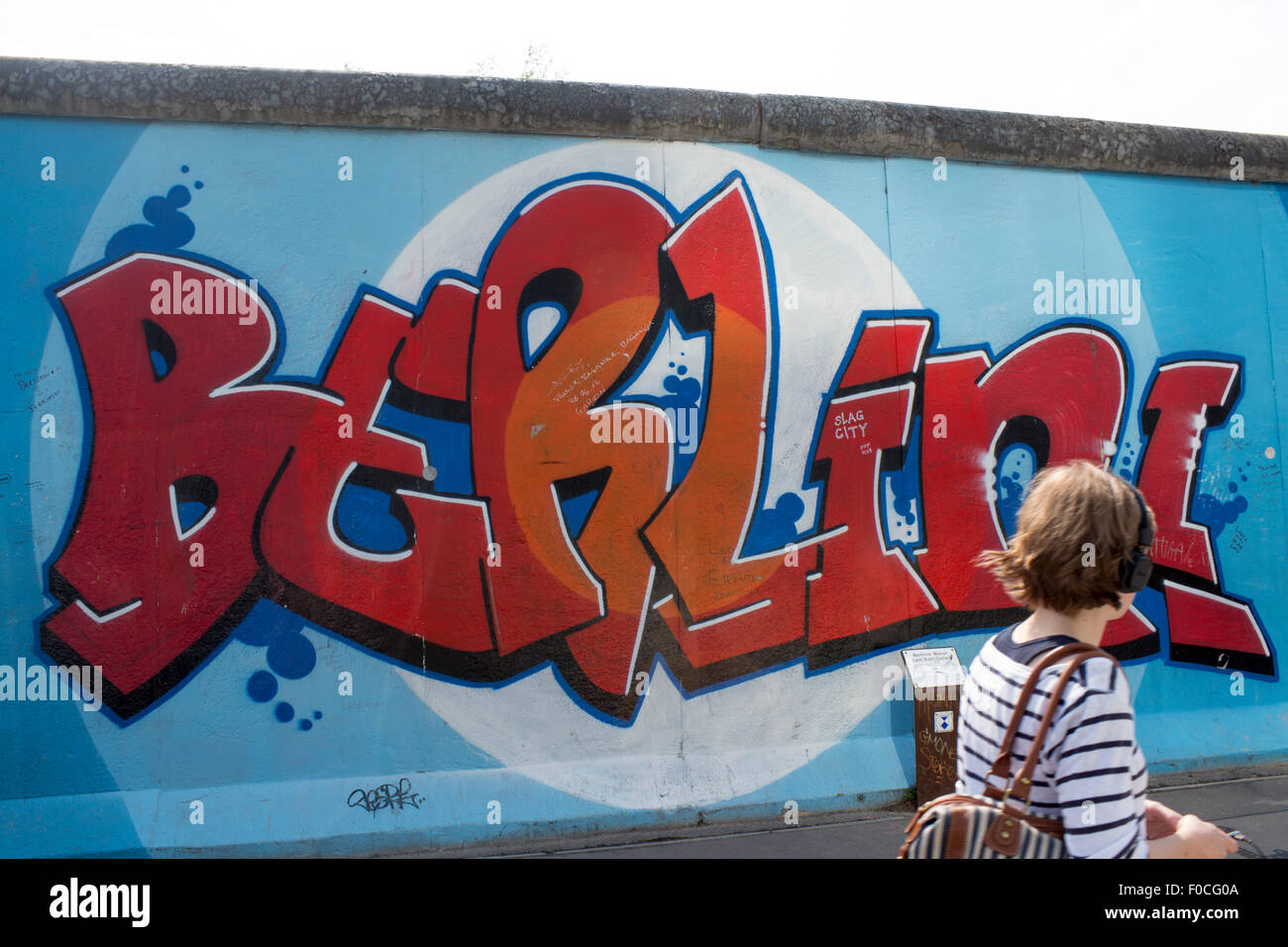 Muro di Berlino East Side Gallery giovane donna persona femmina passato a piedi Berlino! Illustrazione graffiti pittura murale durante l'ascolto Foto Stock