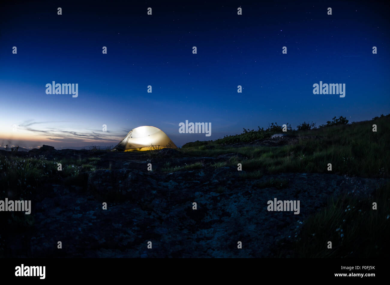 Come il crepuscolo si assesta su una catena montuosa, una tenda è accesa contro un cielo stellato con luci da la valle sottostante ancora incandescente Foto Stock