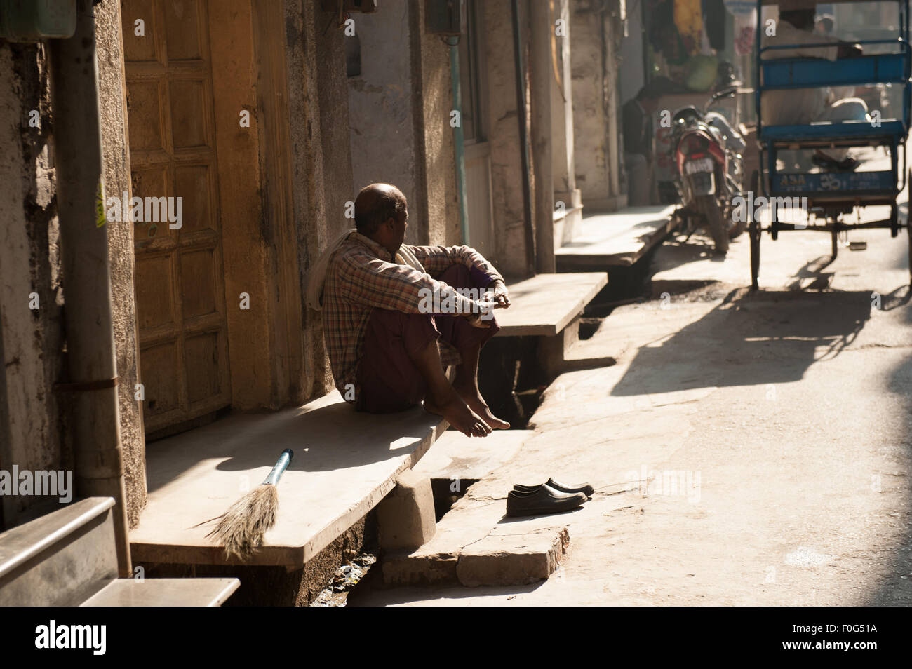 Amritsar Punjab, India. uomo seduto a piedi nudi sul passo della sua casa con un breve spazzare la scopa e le sue scarpe sui gradini. Foto Stock