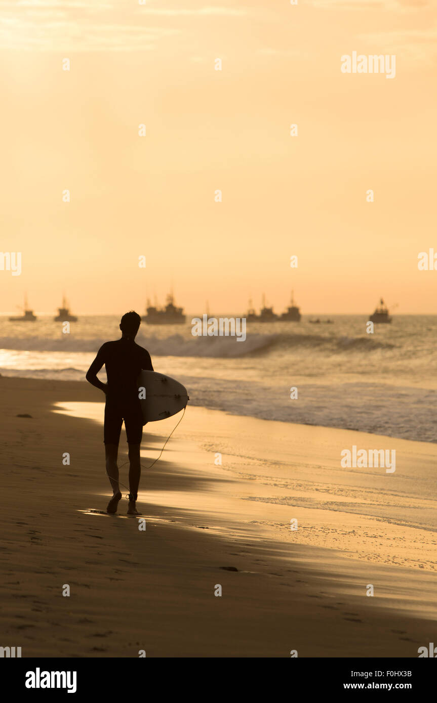 Silhouette solitaria del surfer tenendo la sua scheda e passeggiate sulla spiaggia di Mancora con un giallo arancione tramonto, Perù Foto Stock