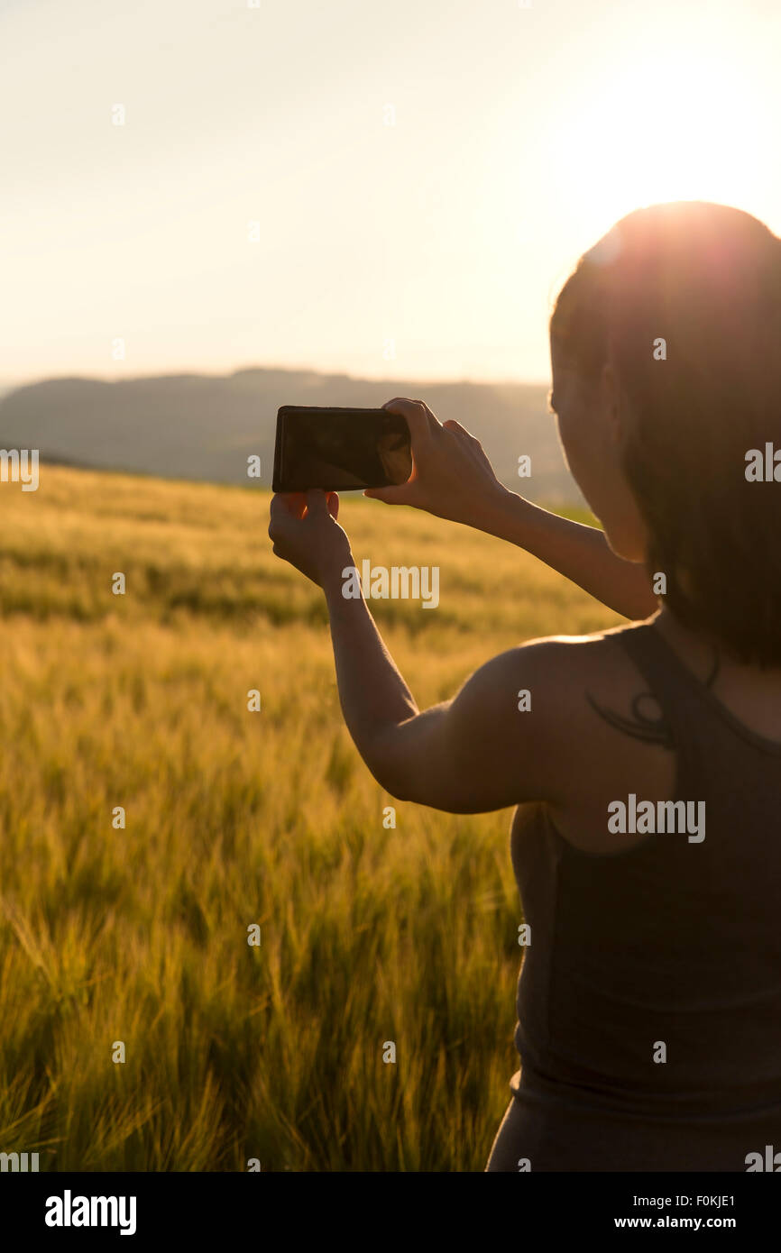Germania, donna in piedi di fronte a un campo a sunrise di scattare una foto con il suo smartphone Foto Stock