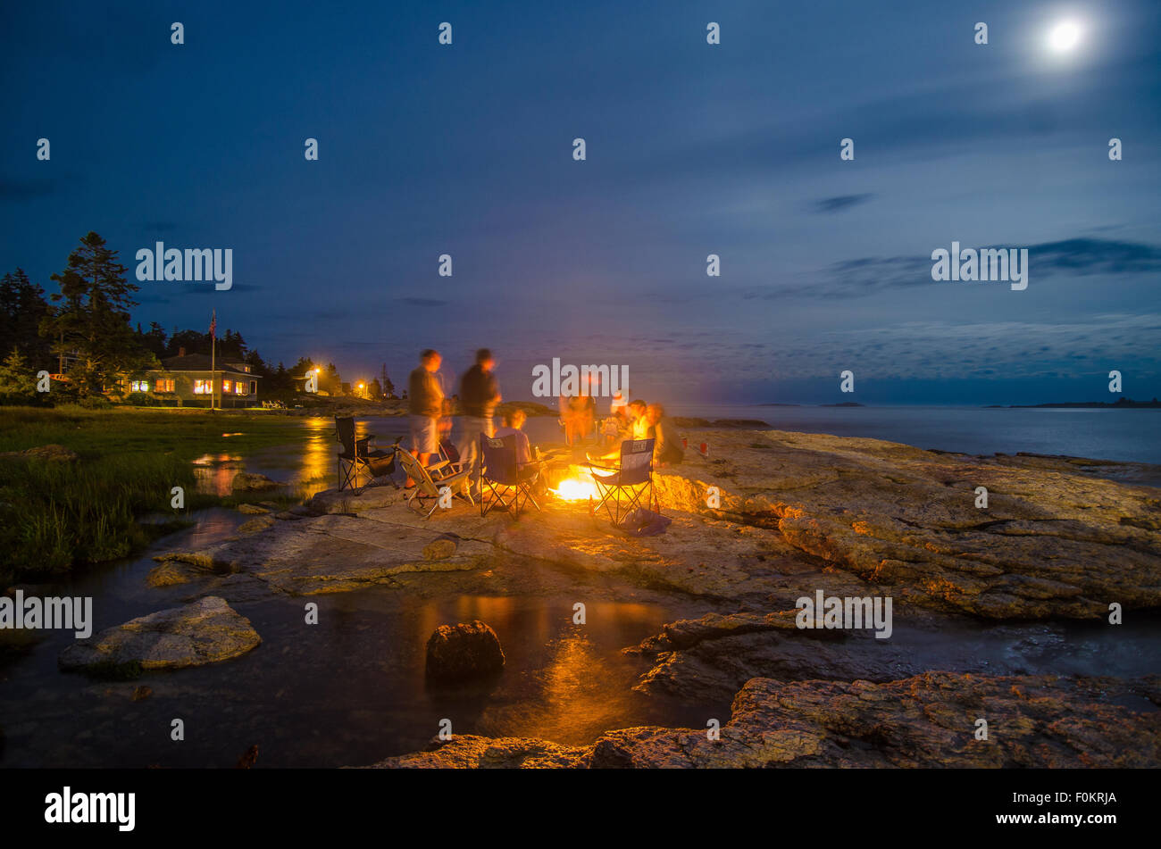 La famiglia e gli amici warm up da un falò sotto una luna illuminata di notte, come alta marea lentamente rivoli entroterra. Foto Stock