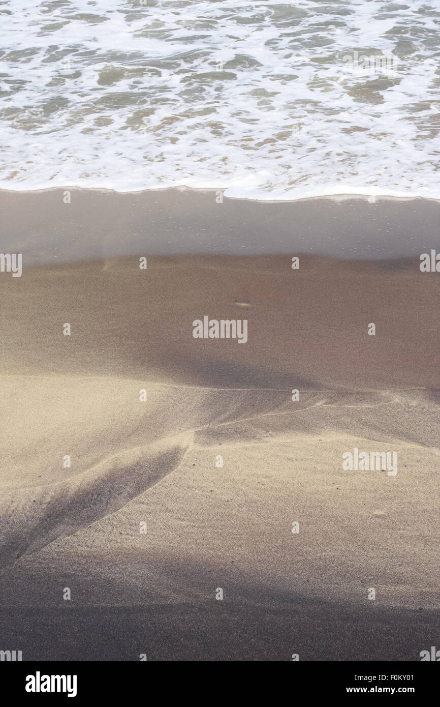 Schiumato in arrivo onde del mare su una spiaggia. Queste onde impetuose lasciare pattern colorati sulla spiaggia con sabbia e limo nero. Foto Stock