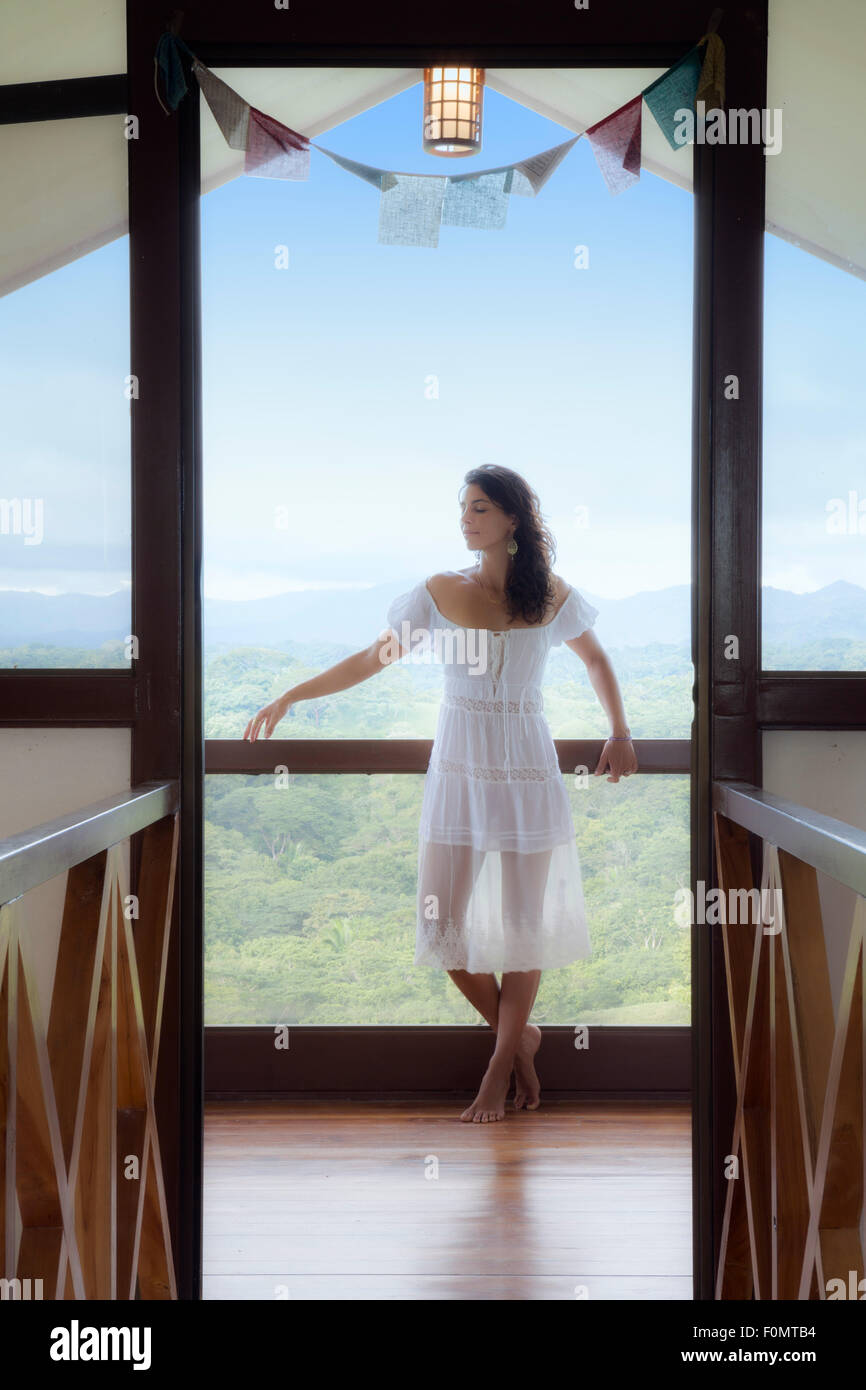 Giovane donna in un vestito bianco in piedi in un meditativo posa e pratica consapevolezza in una cornice porta con blu cielo e foresta dietro di lei Foto Stock