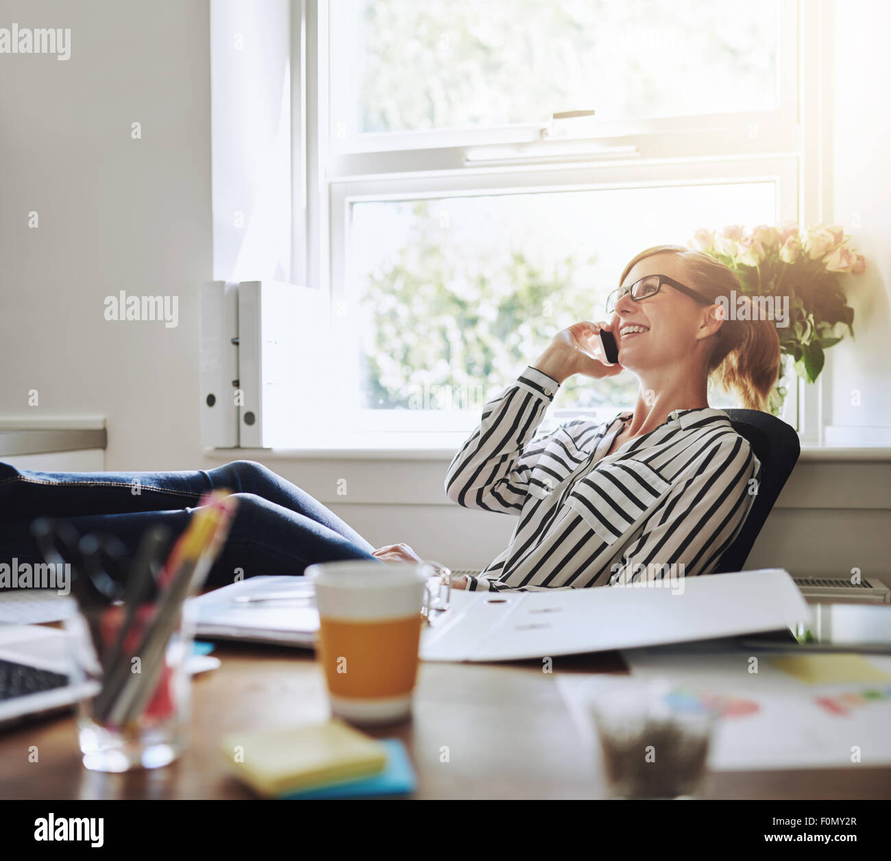 Felice giovane imprenditrice per parlare a qualcuno sul suo telefono cellulare durante i momenti di relax all'interno dell'ufficio con le gambe sulla sua scrivania. Foto Stock