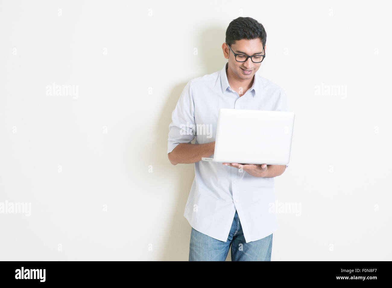 Ritratto di bello business casual uomo indiano utilizzando computer portatile, in piedi su sfondo semplice con ombre, copia spazio a s Foto Stock