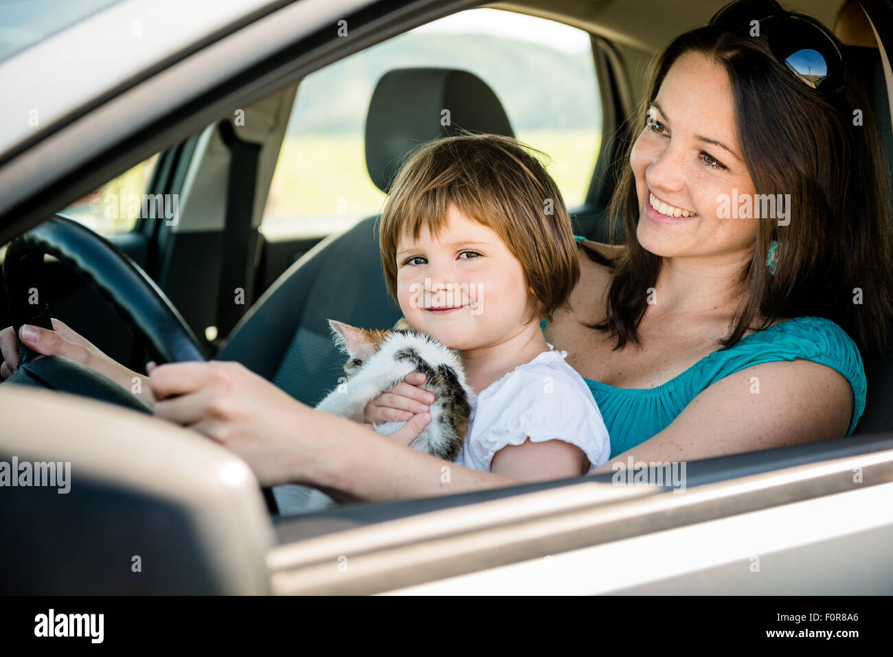 La madre e il Bambino tiene un gattino fingere la guida auto tutti seduti sul sedile anteriore del conducente Foto Stock