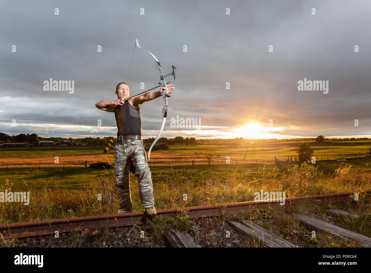 Uomo duro con arco e frecce durante la drammatica NUVOLOSO TRAMONTO. Foto Stock