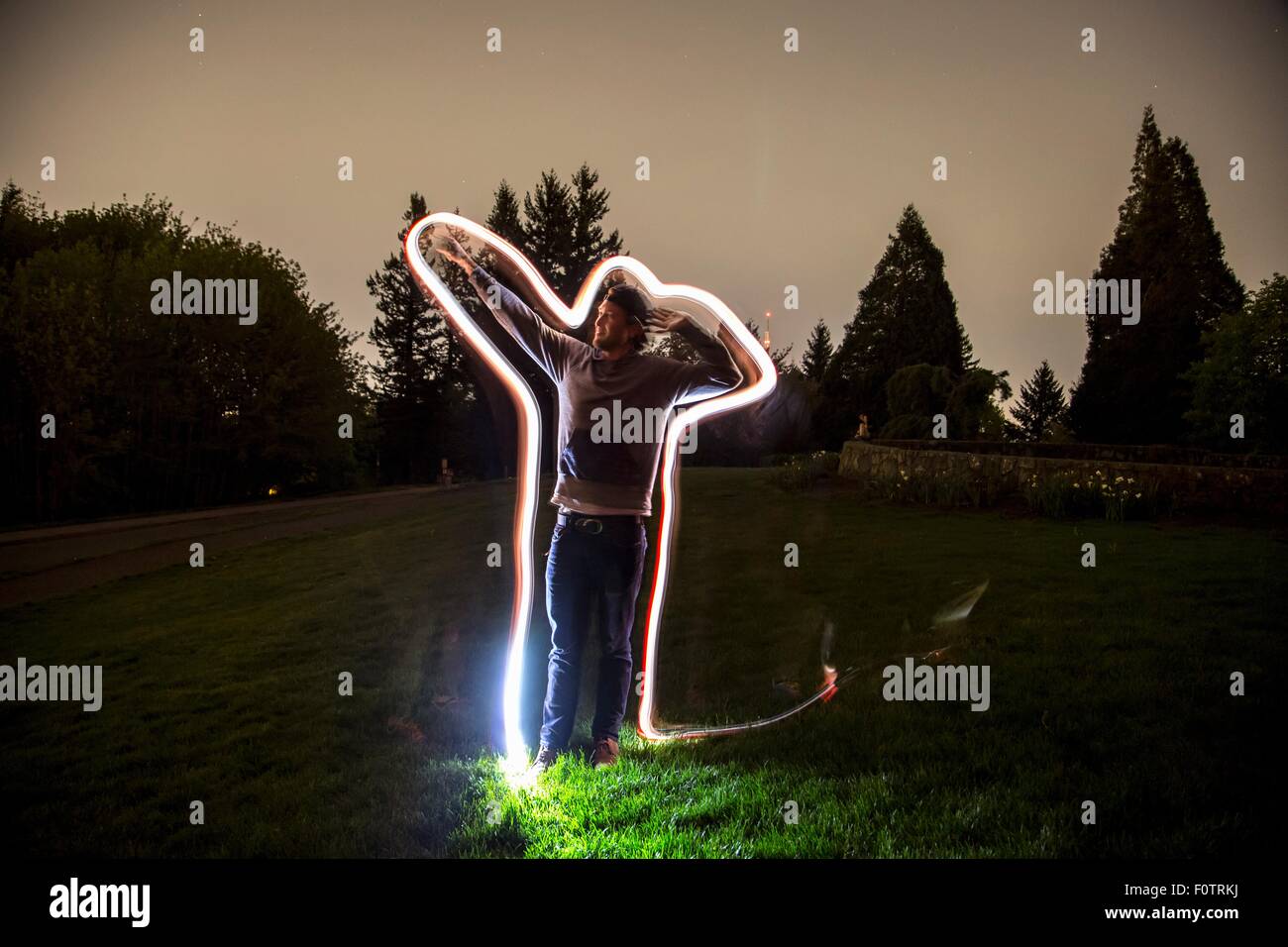 Giovane uomo in piedi in campo al crepuscolo, il braccio in posizione di puntamento, luce sentiero tracing forma del corpo Foto Stock