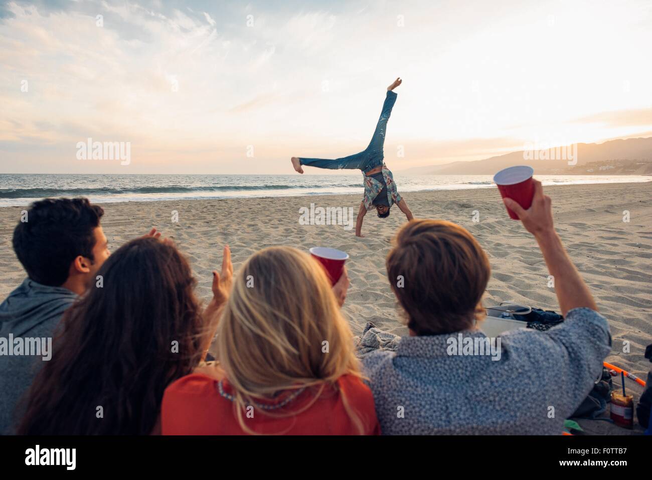 Gruppo di amici seduti sulla spiaggia, guardando amico do cartwheels su e, Tramonto, Vista posteriore Foto Stock