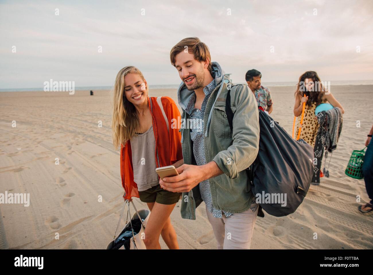 Gruppo di amici a piedi lungo la spiaggia, coppia giovane guardando smartphone Foto Stock