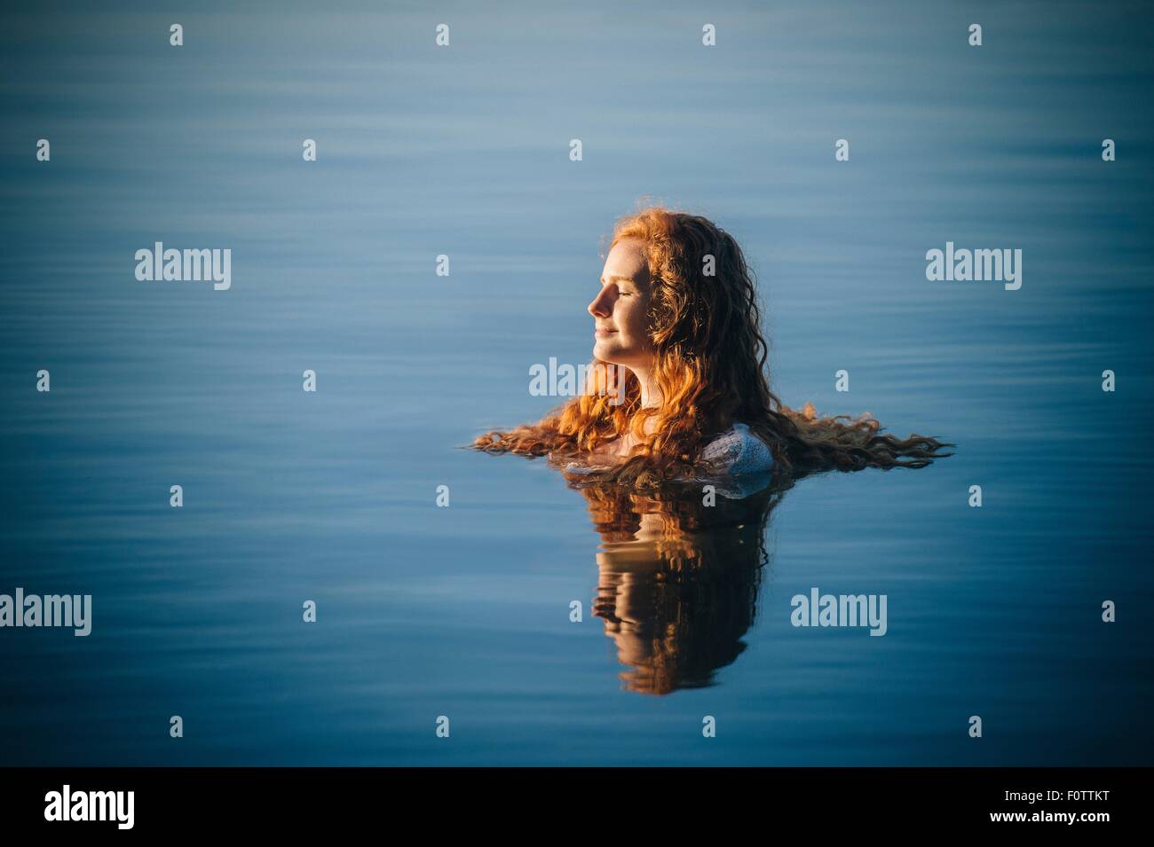 La testa e le spalle del giovane donna con lunghi capelli rossi nel lago con gli occhi chiusi Foto Stock