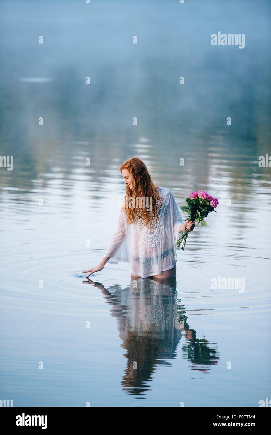 Giovane donna in piedi nel lago holding mazzo di rose e increspa la superficie con le dita Foto Stock