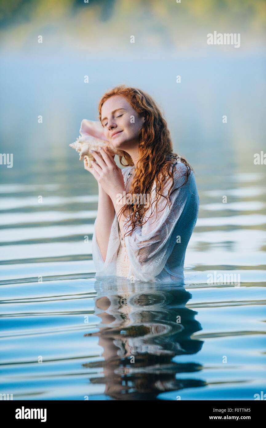 Giovane donna in piedi nel lago misty con gli occhi chiusi ascoltando la conchiglia Foto Stock