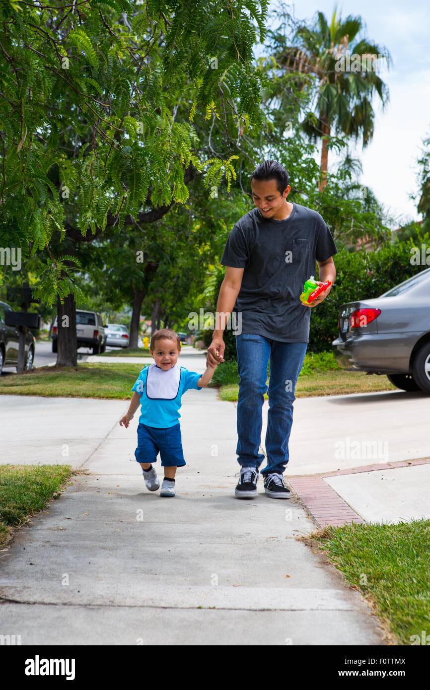 Giovane uomo tenendo le mani con il toddler fratello mentre passeggiando lungo il marciapiede suburbana Foto Stock