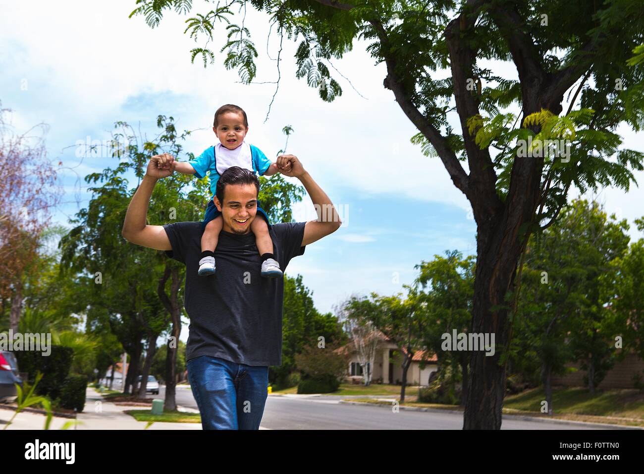 Giovane uomo che porta il toddler fratello su spalle lungo il marciapiede suburbana Foto Stock