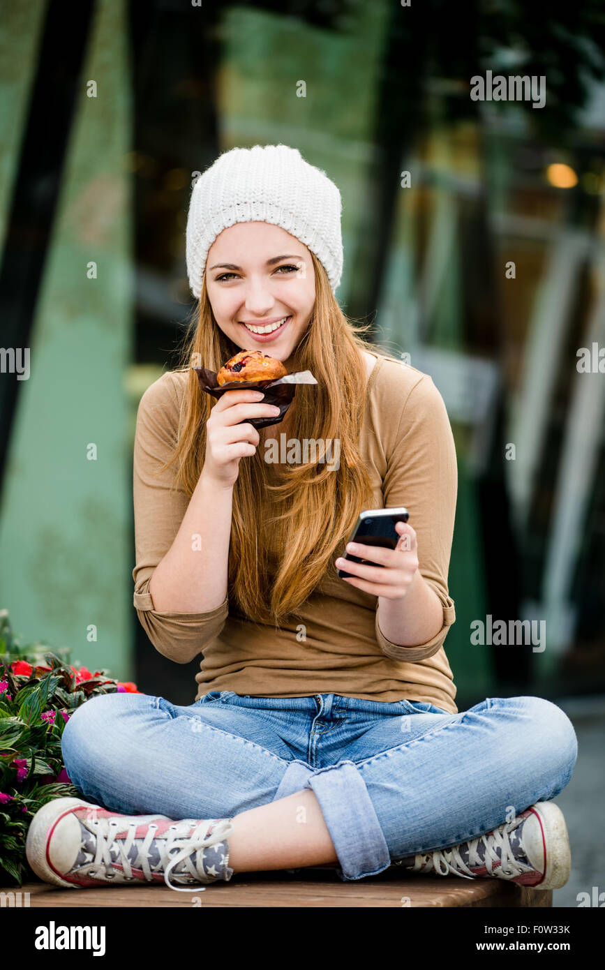 Adolescente - giovane donna mangiare muffin in strada e cercando nel telefono Foto Stock