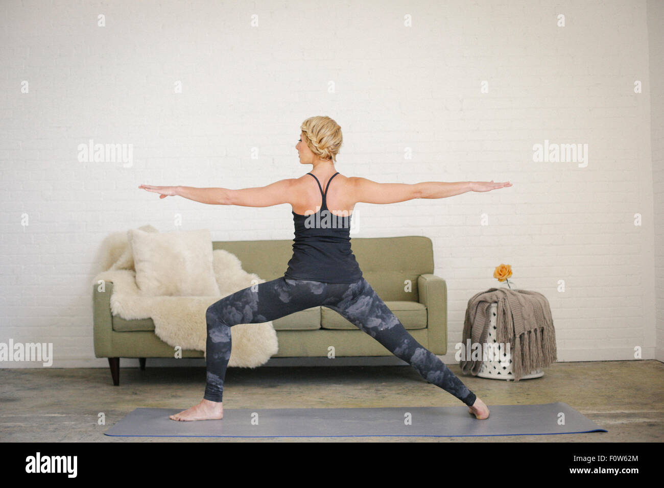 Una donna bionda in un nero leotard e gambali in piedi su un materassino yoga in una stanza, la sua gambe divaricate e braccia tese. Foto Stock